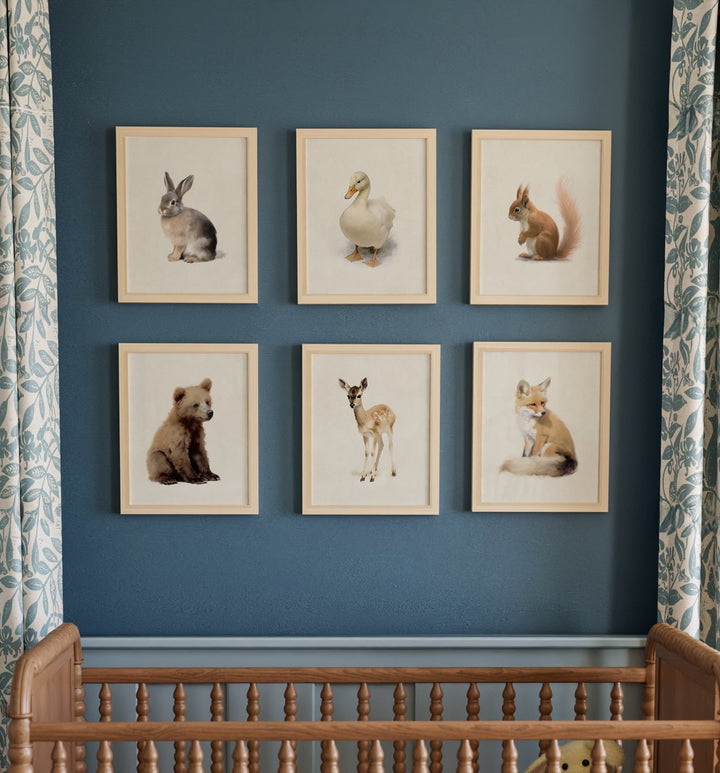 A series of framed prints featuring woodland animal illustrations displayed above a wooden crib in a nursery room, with patterned curtains in the background. The Crown Prints