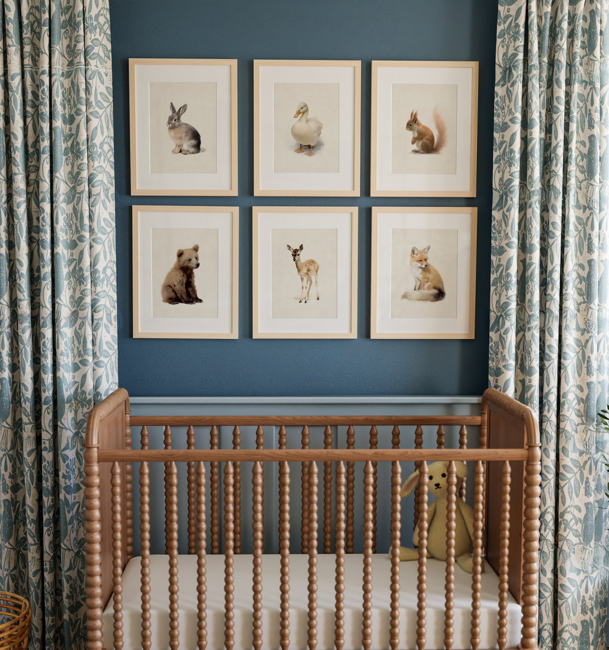 A series of framed prints featuring woodland animal illustrations displayed above a wooden crib in a nursery room, with patterned curtains in the background. The Crown Prints