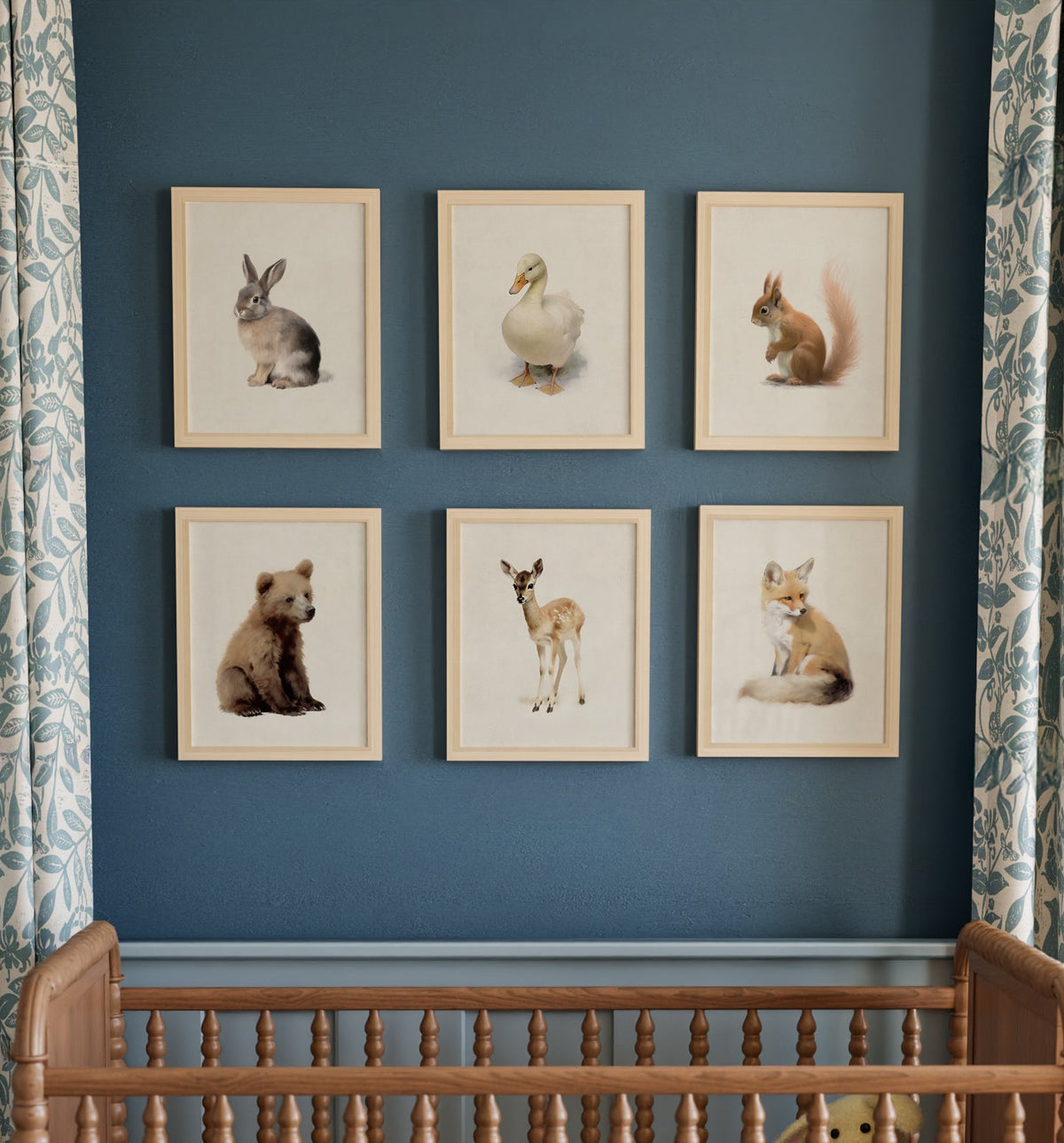 A series of framed prints featuring woodland animal illustrations displayed above a wooden crib in a nursery room, with patterned curtains in the background. The Crown Prints